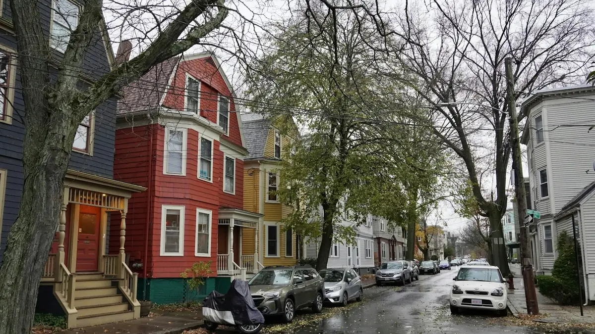 A narrow street of painted multi-family houses with parked cars on a gray day - Leak Detection in Revere